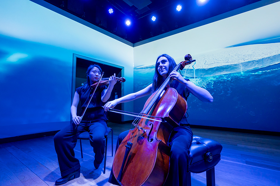 A cellist and violinist performing in a dark space with a photo of a glacier projected around themtest